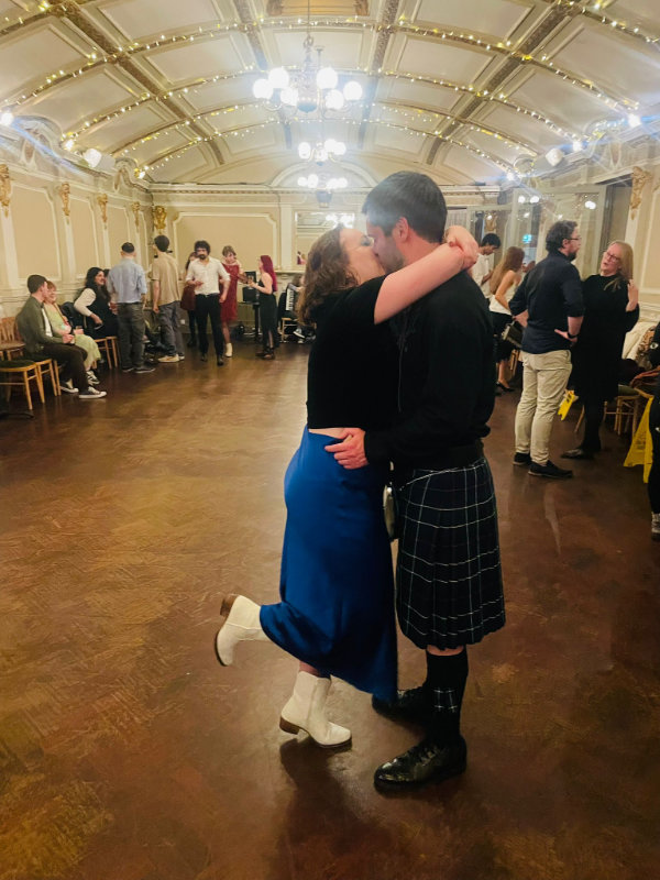Gavin, in a kilt, kissing Amy, in a black top and blue skirt